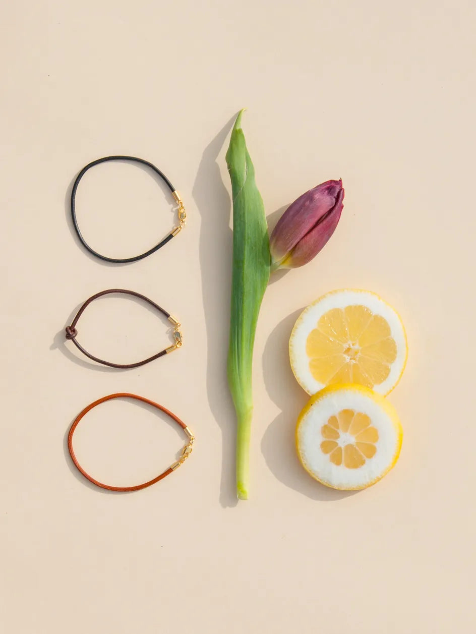 Drei Lederarmbänder in schwarz, dunkelbraun und rotbraun neben einer
Tulpe und 2 Zitronenringen auf cremefarbenem Hintergrund