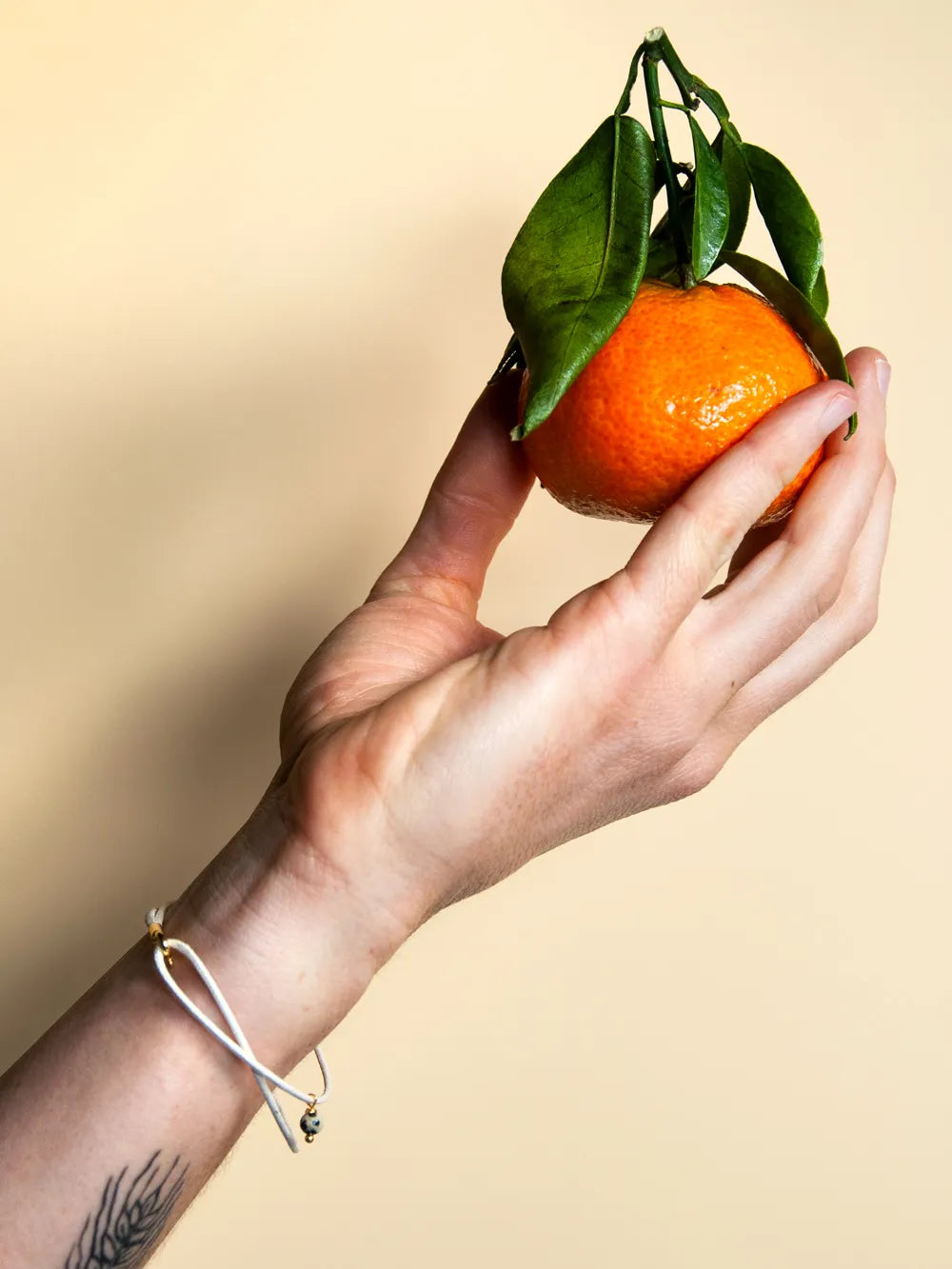 Hand mit cremefarbenem Baumwollarmband mit Steinperle hält eine Mandarine mit Blättern vor gelbem Hintergrund