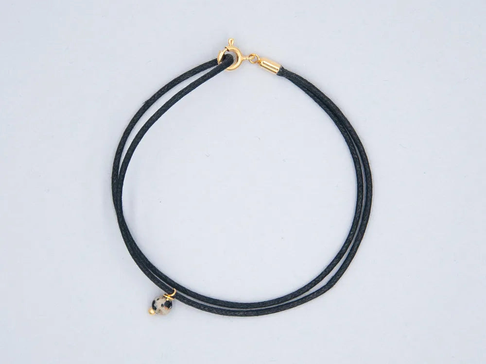 Armband schwarz aus gewachster Baumwollkordel mit runder Steinperle Dalmatinerjaspis und goldenem Verschluss vor weissem Hintergrund