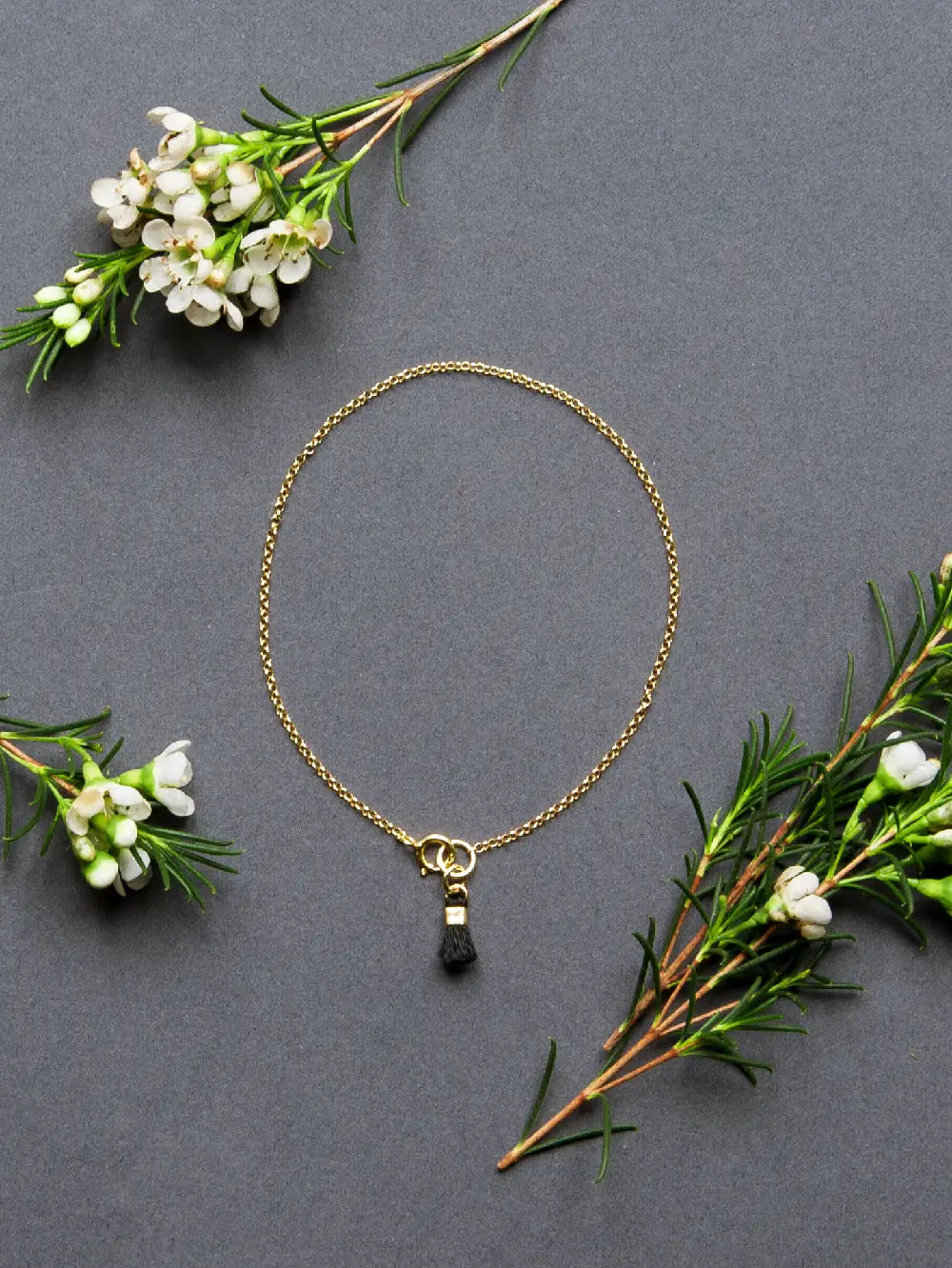 Armband aus goldener Kette mit schwarzem Quastenanhänger, deneben 3 grüne Zweige mit weissen Blüten auf grauem Hintergrund