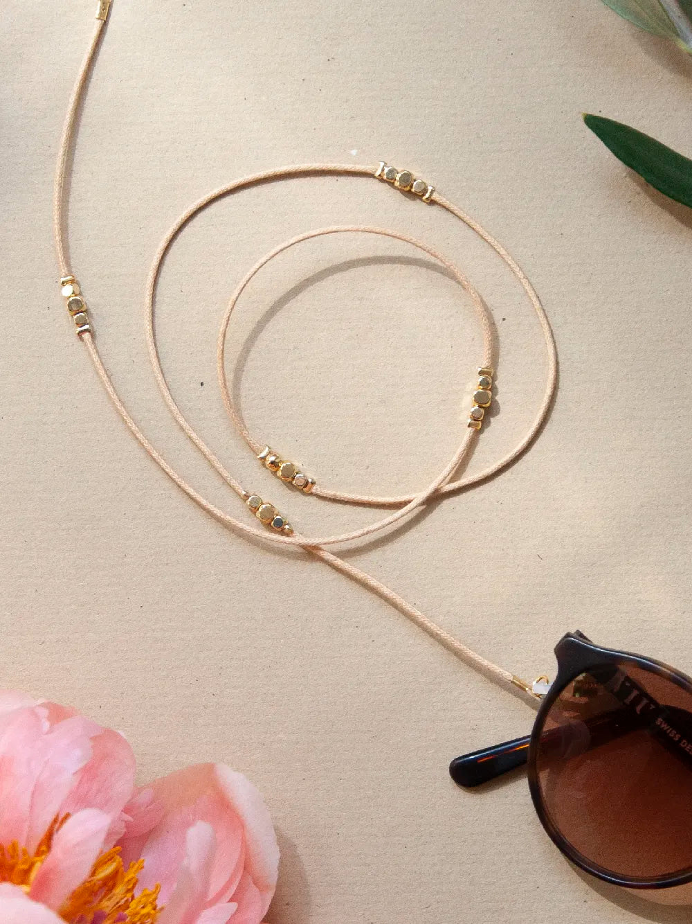 Brillenband crème aus gewachster Baumwollkordel in 1,5 mm Dicke mit goldenen Perlen an Sonnenbrille auf beigem Hintergrung