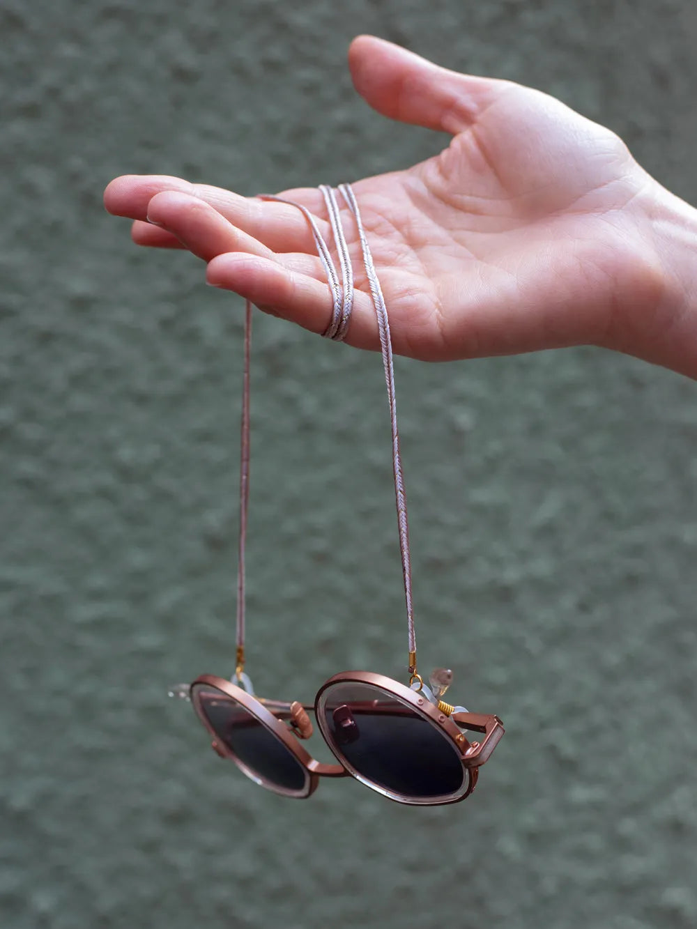 Hand mit umgewickeltem cremefarbenen Brillenband an Sonnenbrille vor olivfarbenem Hintergrund