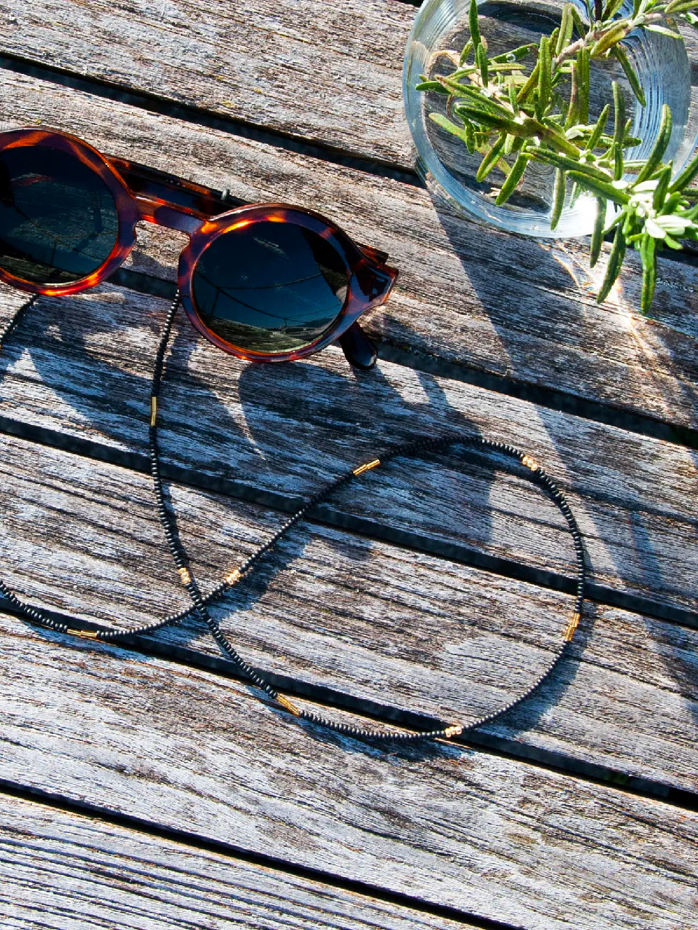 Brillenband aus schwarzen Glasperlen mit goldenen Akzenten an Sonnenbrille auf Holzlatten, Glas mit Rosmarin daneben