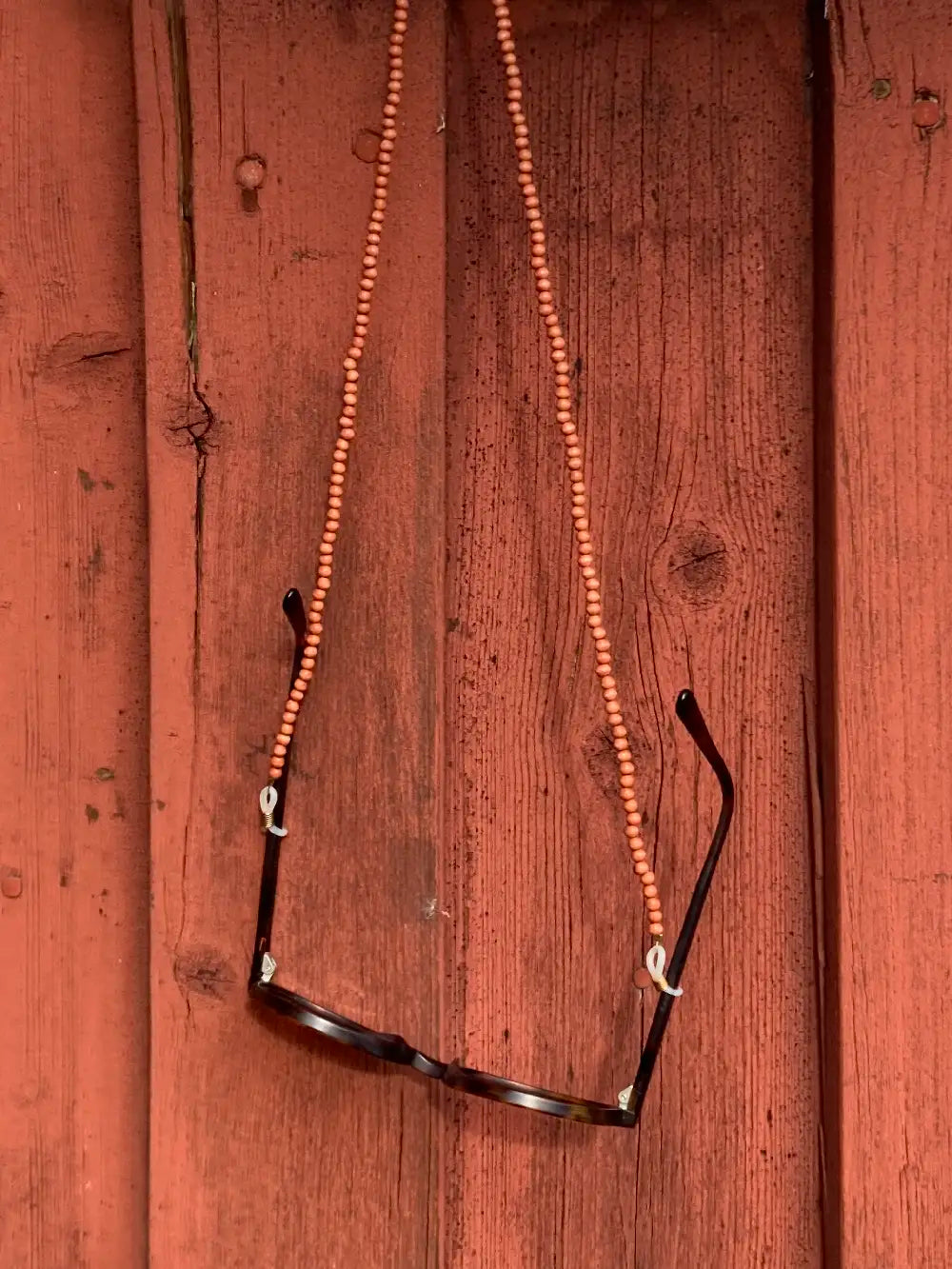 Eine Sonnenbrille hängt an einem Brillenband aus hellbraunen Holzperlen vor einer brauenen Holzwand