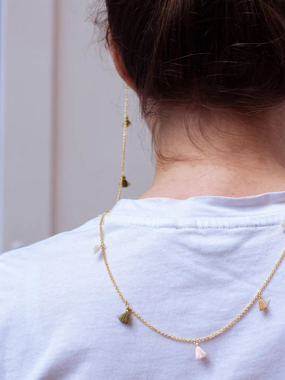 Nacken einer Person mit braunem Haar und weissem T-Shirt, goldenes Brillenband aus Kette mit creme, beige und oliv Quasten fällt auf den Rücken