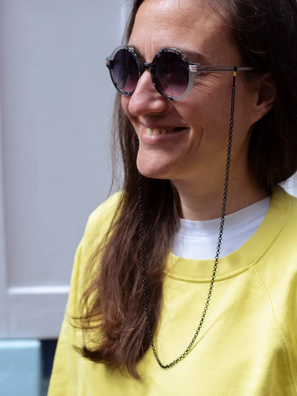 Eine Person mit langen braunen Haaren und gelbem Pulli trägt eine runde Sonnenbrille mit einem Brillenband aus schwarzer Kette