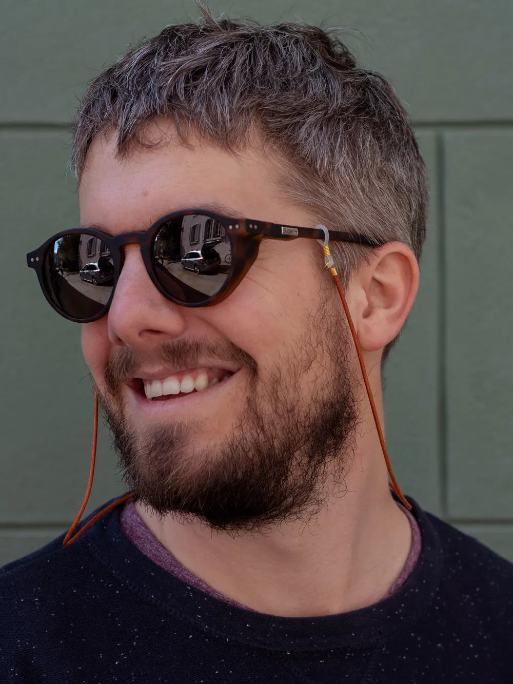 Mann mit grauen Haaren trägt Sonnenbrille mit Brillenband aus rotbraunem Leder vopr olivgrünem Hintergrund