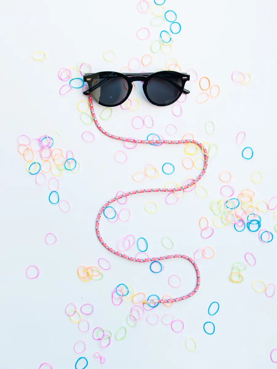 Pinkes Brillenband an schwarzer Sonnebrille auf weissem Hintergrund mit vielen farbigen Gummibändern