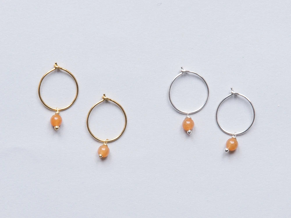 Zwei Paar Creolen in gold und silber mit je einer orangen runden Steinperlen auf grauem Hintergrund