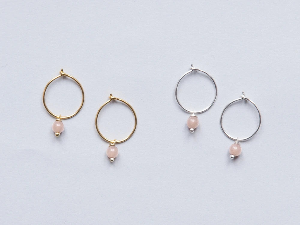 Zwei Paar Creolen in gold und silber mit je einer rosa runden Steinperlen auf grauem Hintergrund
