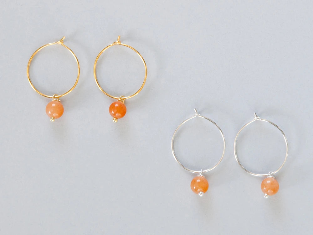 Zwei paar Creolen, einmal in silber und einmal in gold mit je einem runden Steinanhänger in orange auf weissem Hintergrund