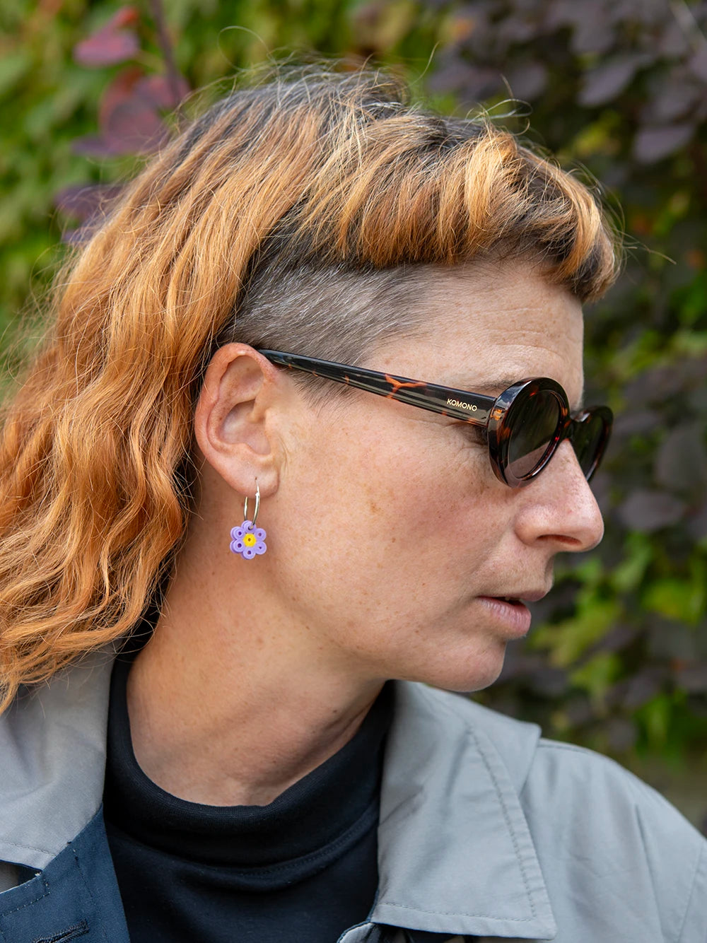 Seitliche Ansicht einer Frau mit orangen Haaren die eine Sonnenbrille trägt. Die Frau trägt silberne Creolen mit einem Anhänger aus lila farbenen Bügelperlen.