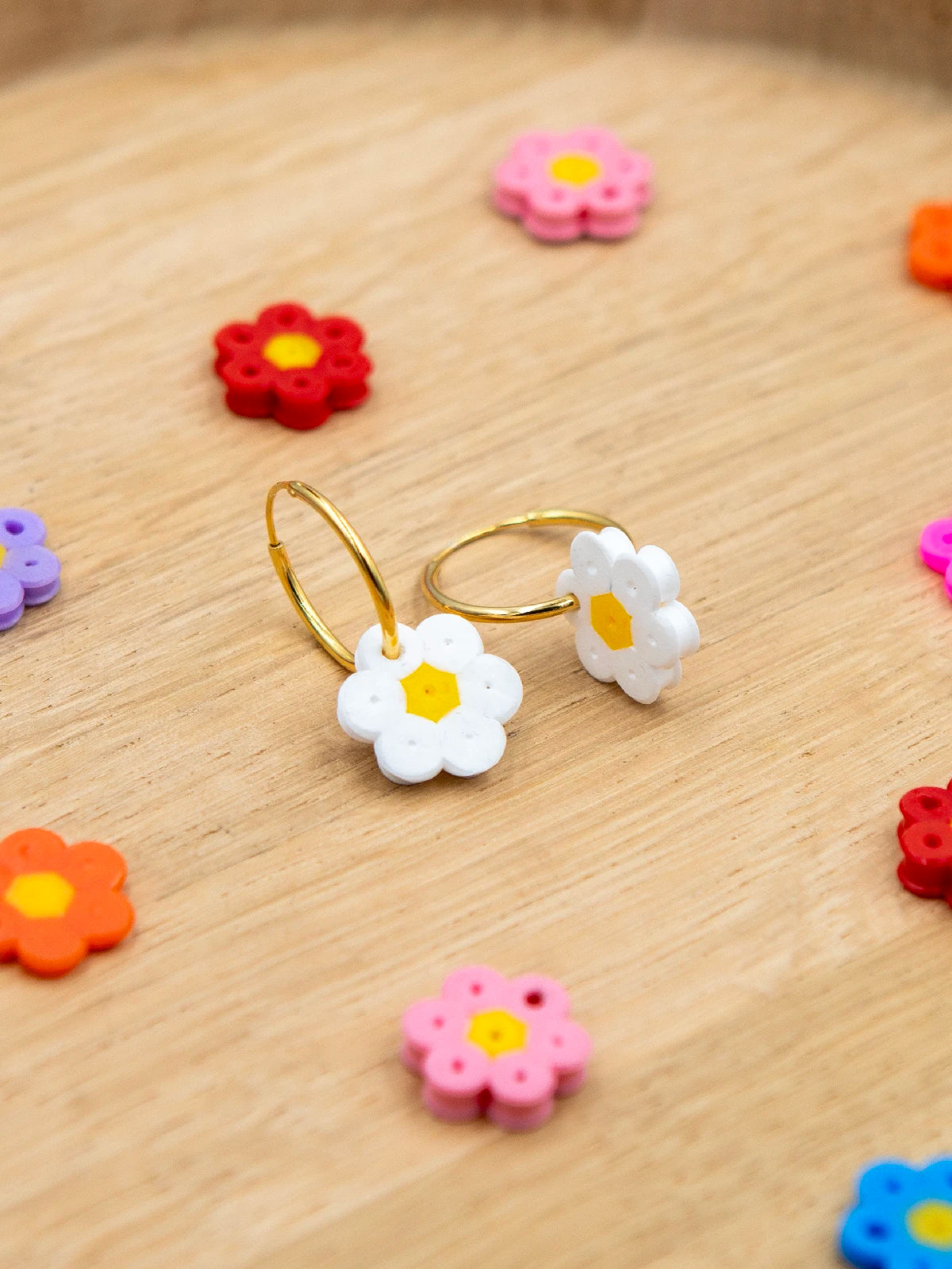Zwei goldene Creolen mit Blume aus weissen Bügelperlen als Anhänger und weiteren solchen Blumen auf holzigem Untergrund