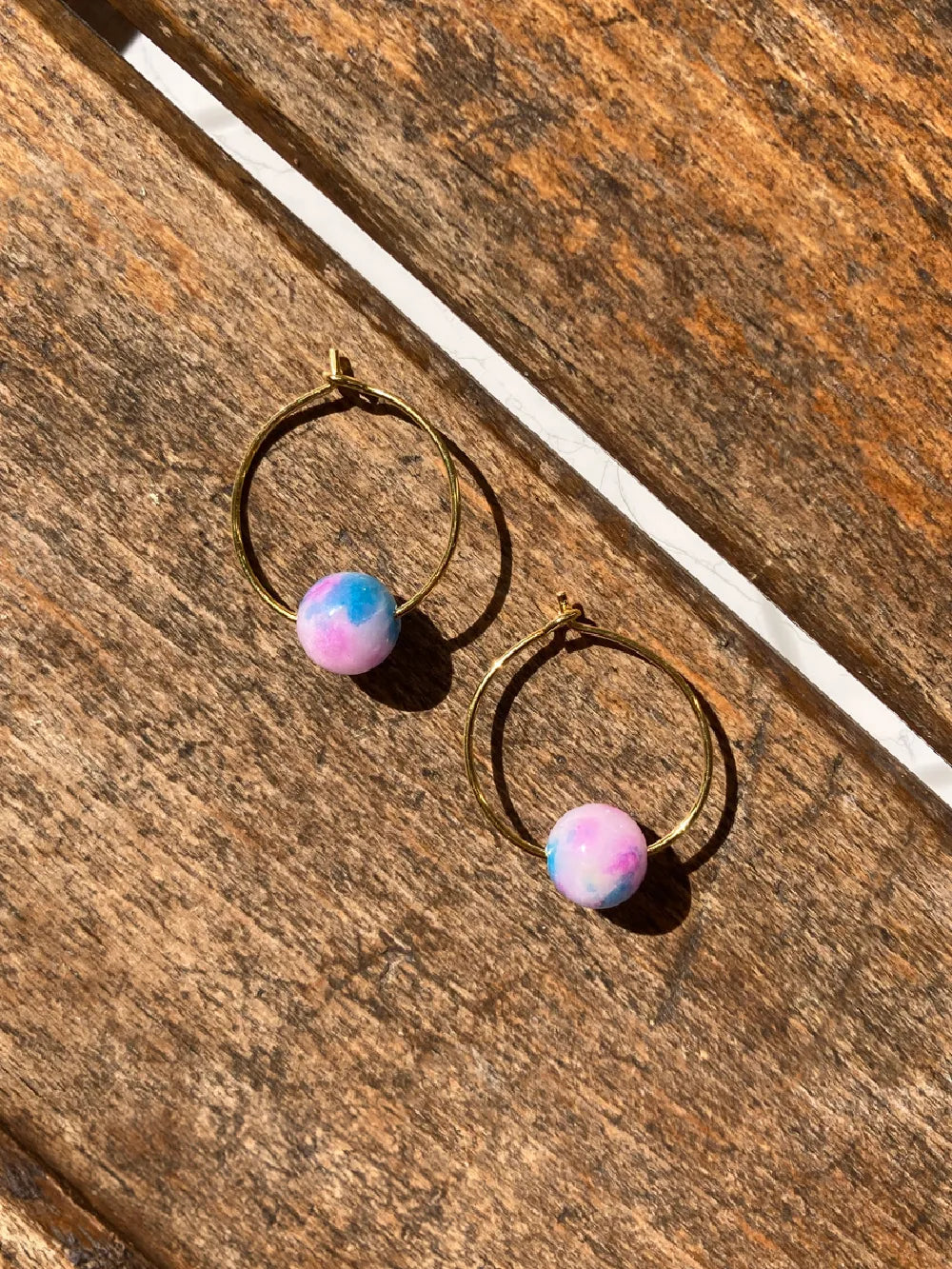 Zwei goldene Creolen mit je einer runden Steinperle in pink, blau und weiss auf brauner Holzunterlage