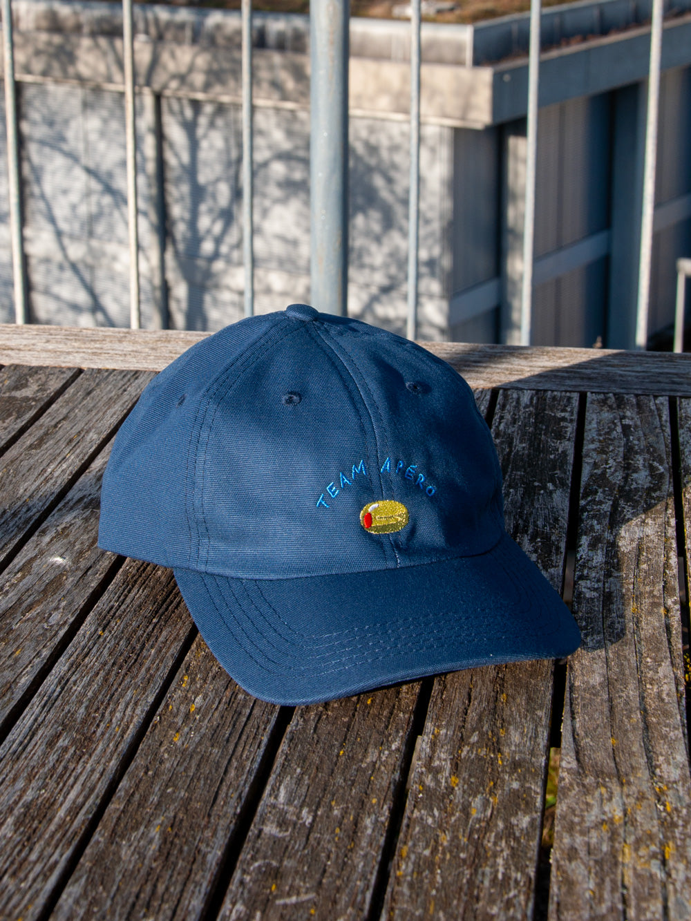 Dunkelblaues Dad Cap mit der Stickerei Team Apéro auf verwittertem Holztisch mit Gebäude im Hintergrund.