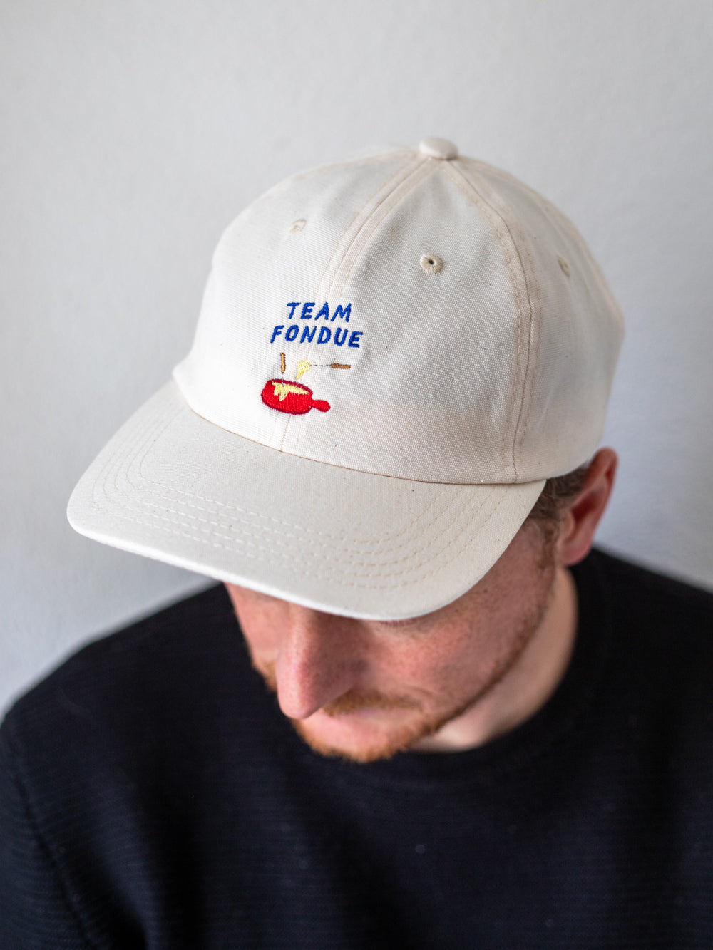 Oberkörper eines Mannes, der ein ecru farbenes Cap und einen schwarzen Pullover trägt. Darauf ist eine Stickerei mit dem Schriftzug Team Fondue und der Abbildung eines roten Fondue Caquelons.