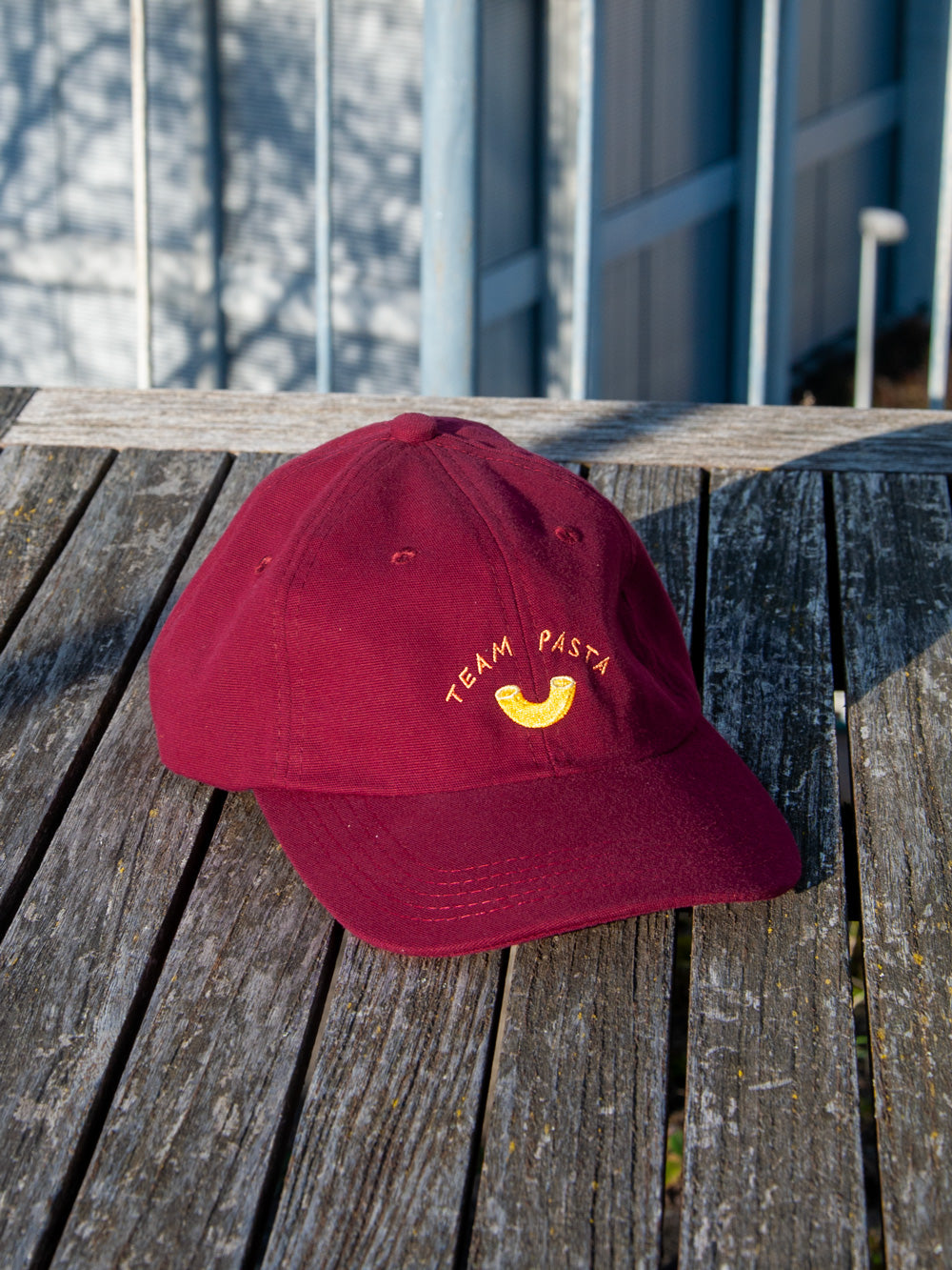 Weinrotes Dad Cap mit der Stickerei Team Pasta und einem ebenfalls gestickten gelben Hörnli auf verwittertem Holztisch und mit Gebäuden im Hintergrund.