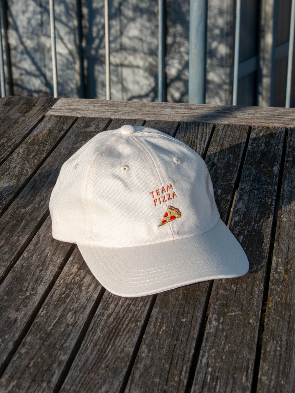 Nature farbenes Dad Cap mit der Stickerei Team Pizza auf verwittertem Tisch mit Gebäuden im Hintergrund