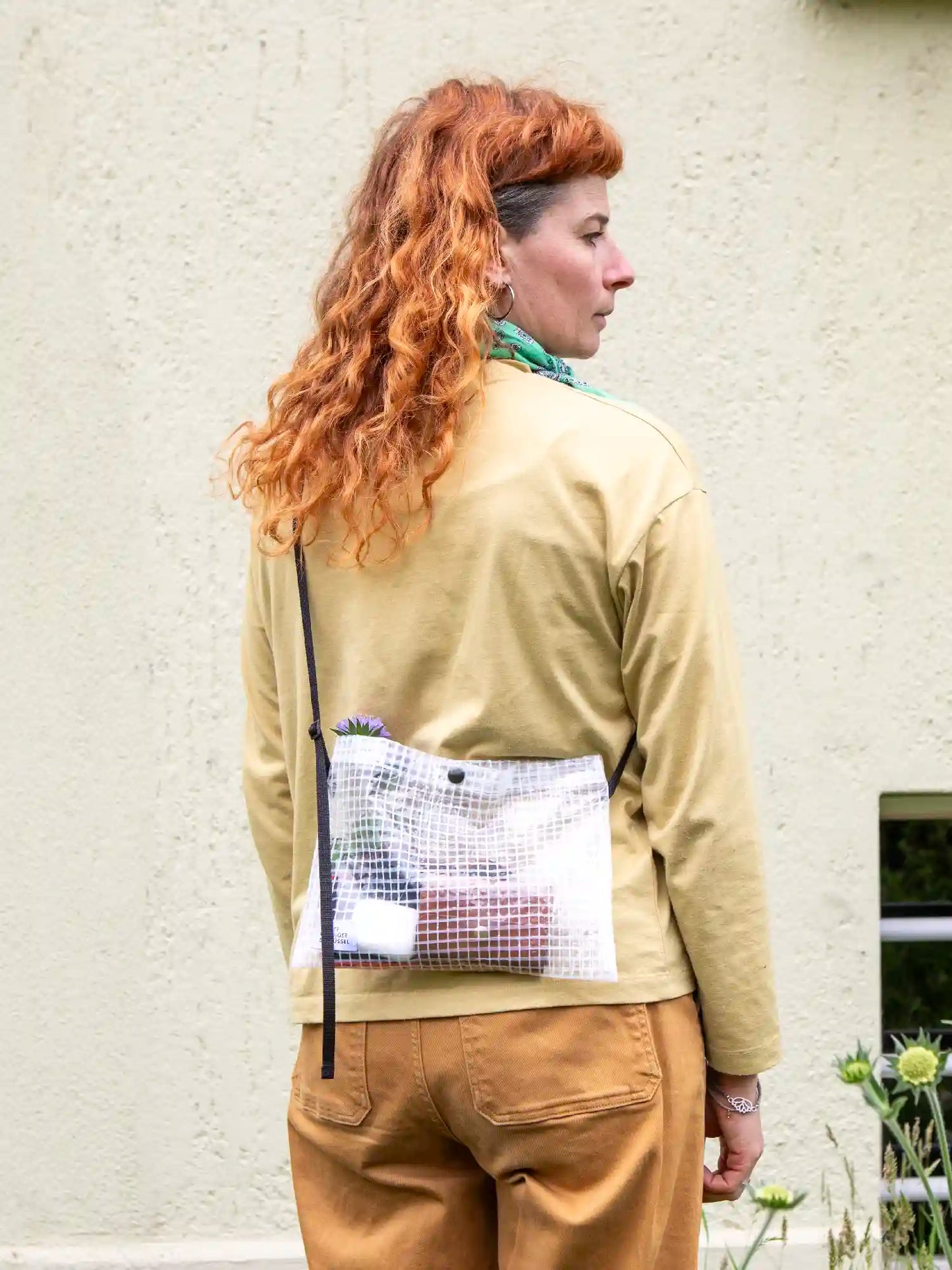 Frau mit langen orangen Haaren und Pony trägt eine Umhängetasche aus transparentem Material mit Gittermuster. Die Umhängetasche ist gefüllt mit einem Portemonnaie, AirPods und einer violetten Blume. Im Hintergrund sieht man eine gelbgrüne Hausfassade.