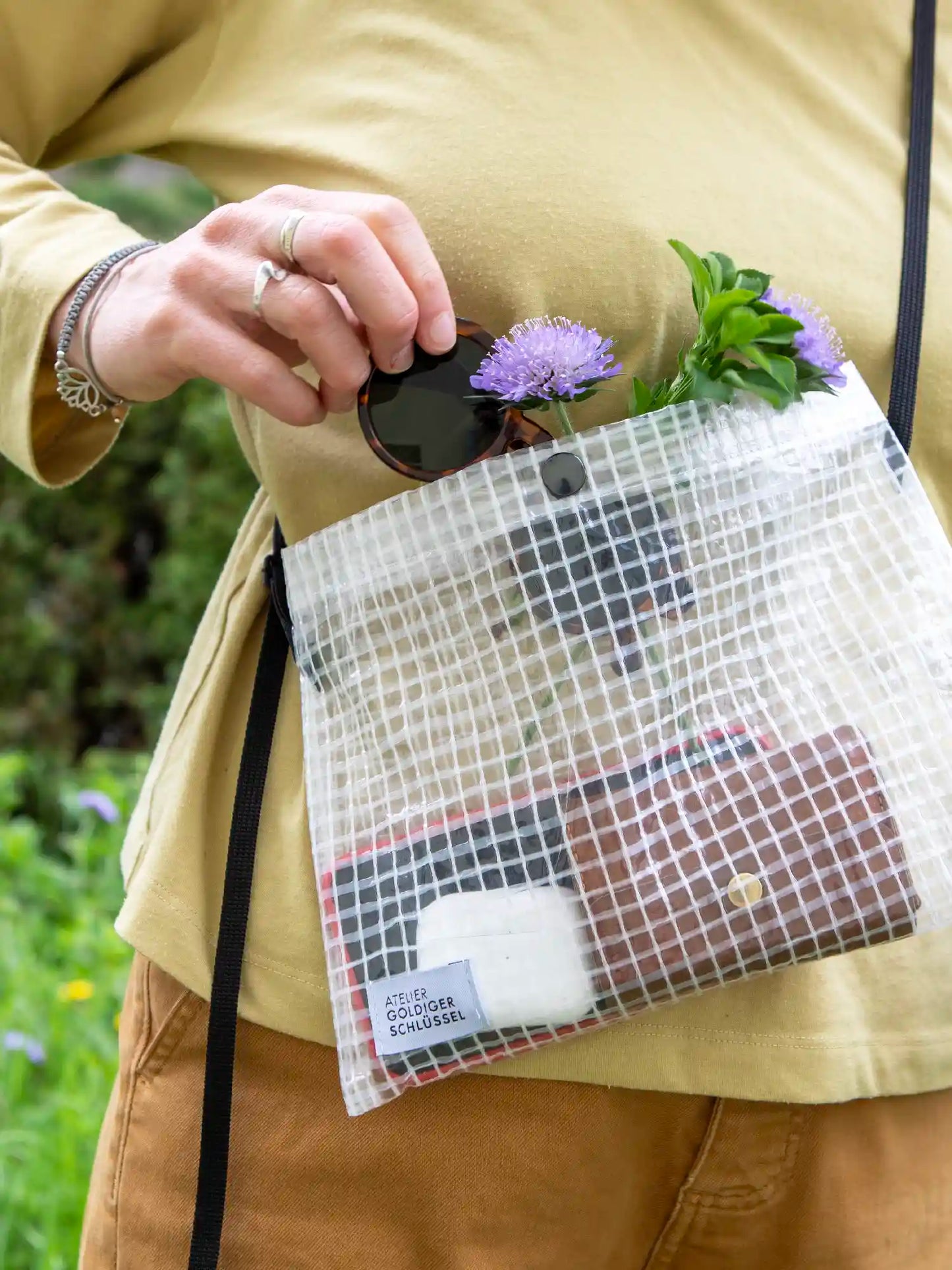 Detailaufnahme einer Musette aus transparentem Material mit Gittermuster. Die Umhängetasche mit scharzen Bändern ist gefüllt mit einem Portemonnaie, AirPods und einer Sonnernbrille. Die Person welche die Tasche trägt ist in gelb braun Tönen angezogen.