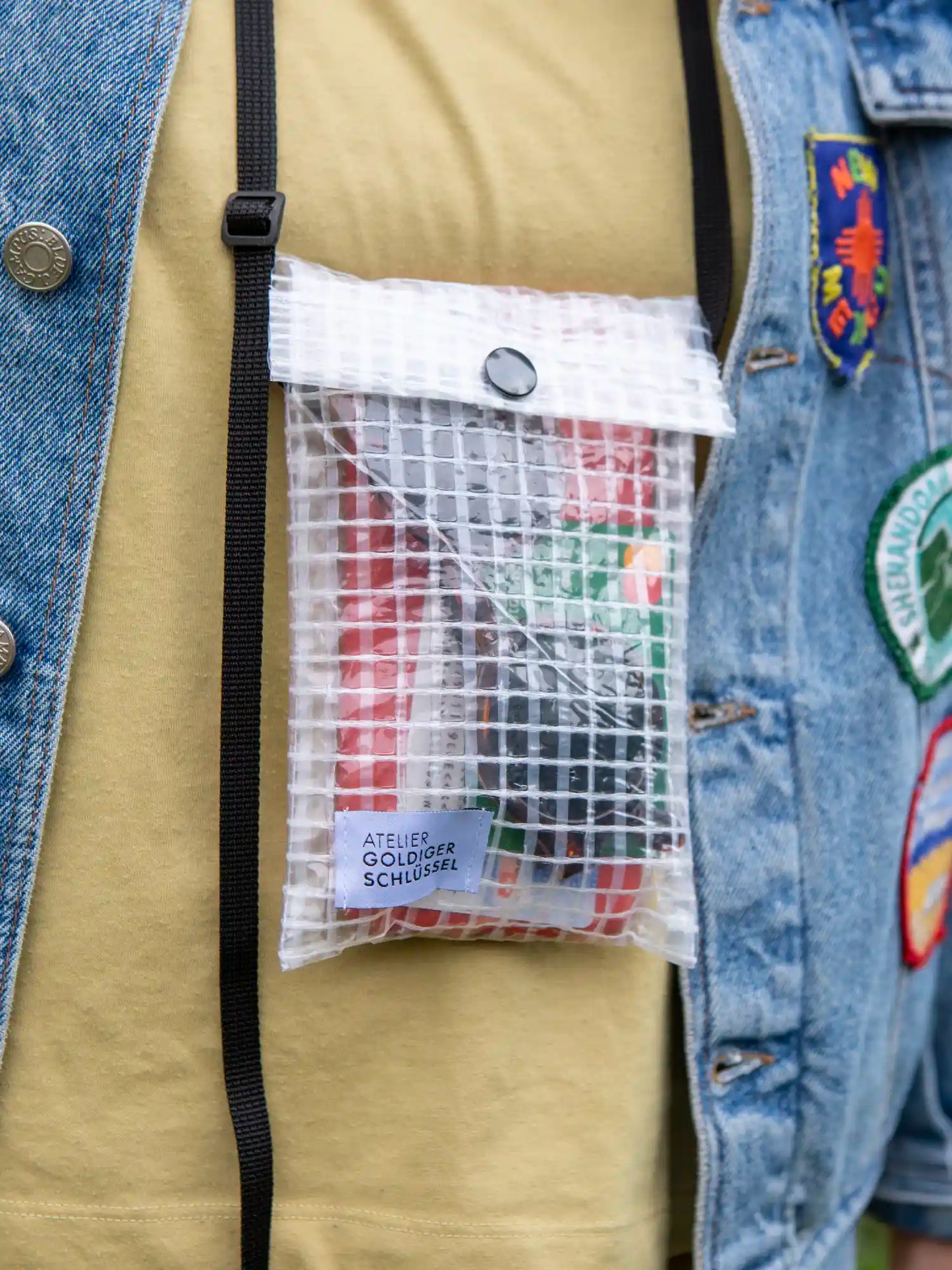 Detailaufnahme einer kleinen Tasche mit schwarzem Druckknopf und Bändern die aus einem transparenten wasserfesten Material mit Gittermuster besteht. Im Innern der Tasche sieht man einen Sonnenbrille und eine Bankkarte. Die Tasche ist mit einem Stofflabel von "Atelier Goldiger Schlüssel" bestückt.