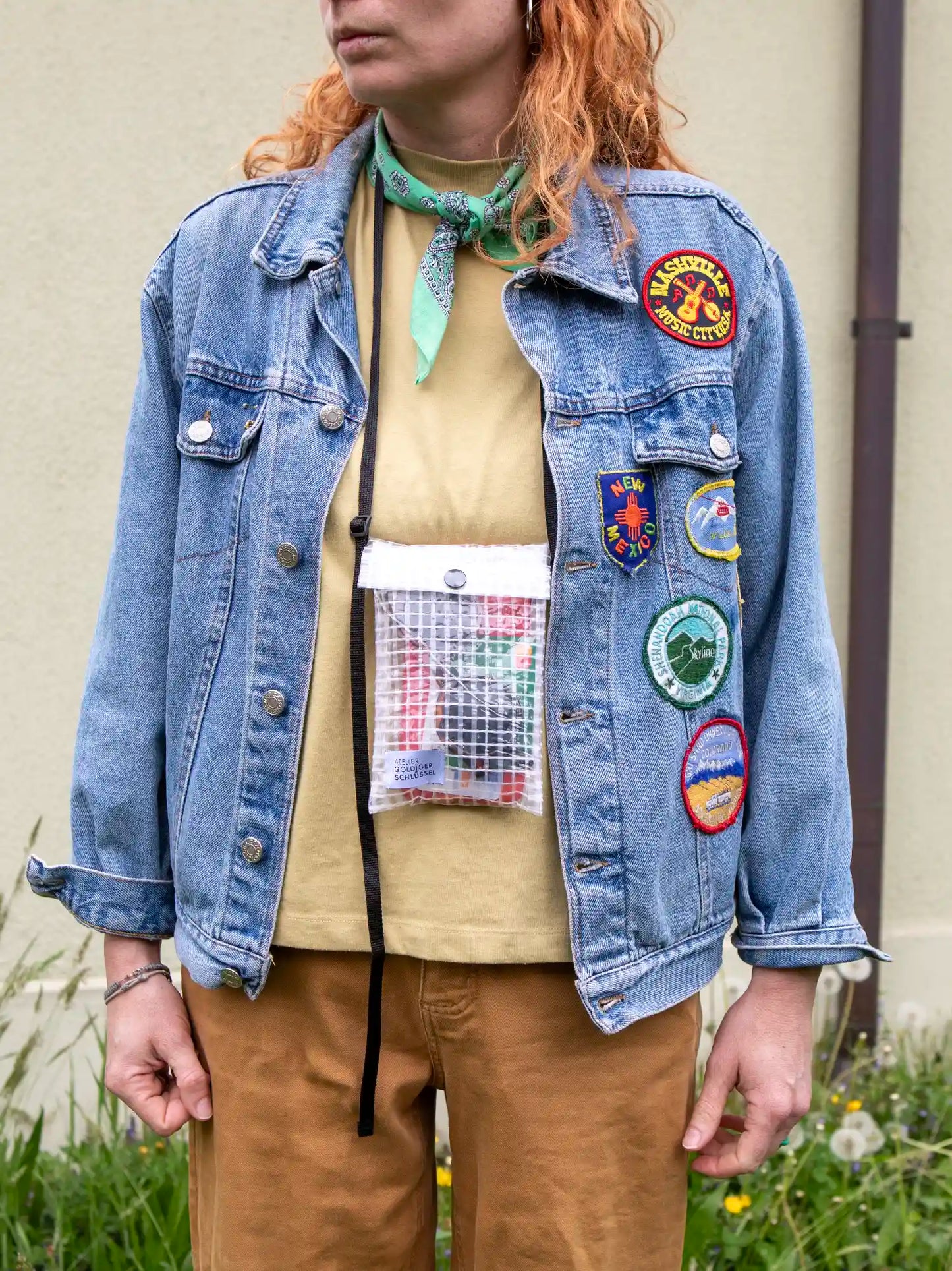 Frau mit langen orangen Haaren trägt eine kleine Tasche aus transparentem Material mit Gittermuster um den Hals. Die Umhängetasche ist gefüllt mit einer Sonnenbrille und einer Bankkarte. Die Frau trägt eine Jeansjacke mit bunten Patches, im Hintergrund sieht man eine gelbgrüne Hausfassade.