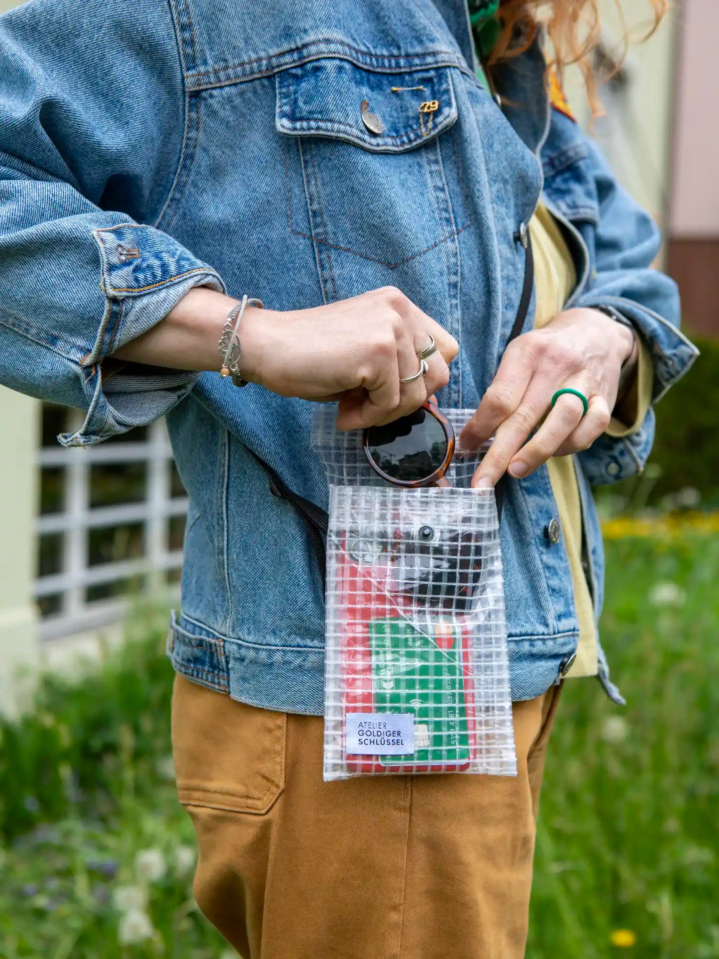 Oberkörper einer Person mit blauer Jeansjacke die sich eine kleine Tasche mit Druckknopfverschluss ungehängt hat. Die Person zieht gerade eine Sonnenbrille aus der Tasche welche ausserdem mit einer Bankkarte und einem iPhone gefüllt ist. Die Tasche hat das Label "Atelier Goldiger Schlüssel". Im Hintergrund sieht man eine Wiese.