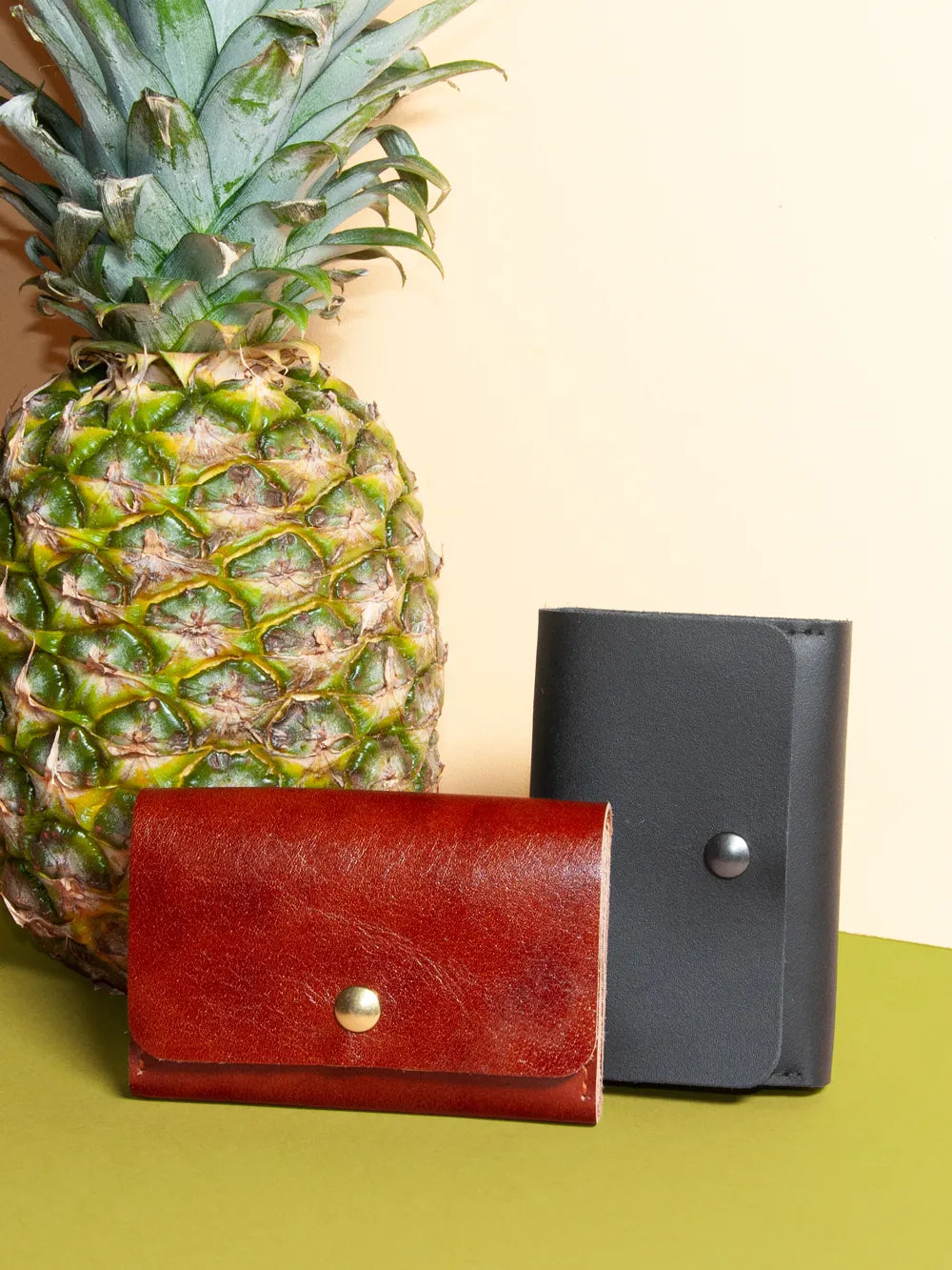 Zwei Portemonnaies aus Leder stehen vor einer Ananas auf einer grünen Fläche