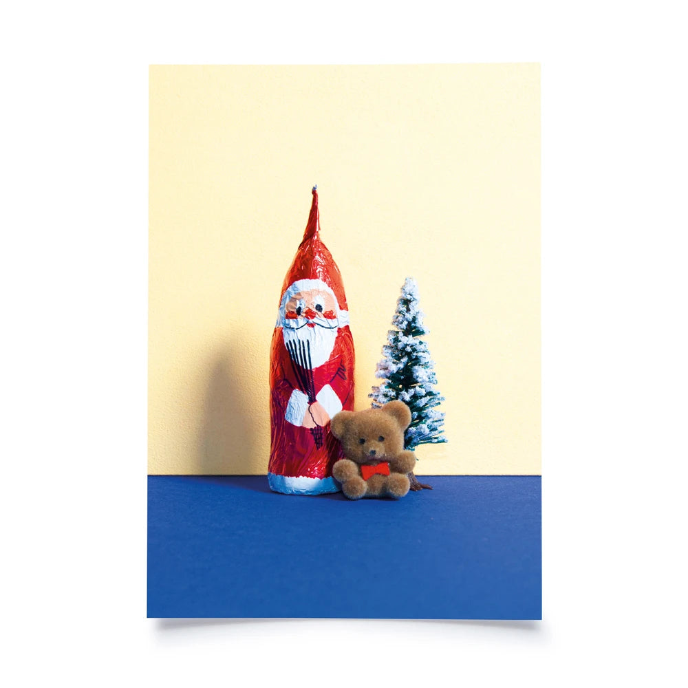Postkarte mit Farbflächen in creme und blau als Hintergrund, davor ein Schokosamichlaus,daneben eine Plastiktanne und ein kleiner Teddybär
