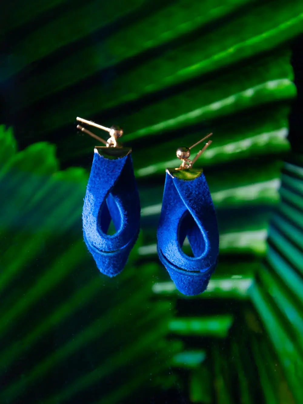 Ohrhänger aus blauem Recycling Leder mit goldenen Metallteilen auf Hintergrund aus Palmblättern