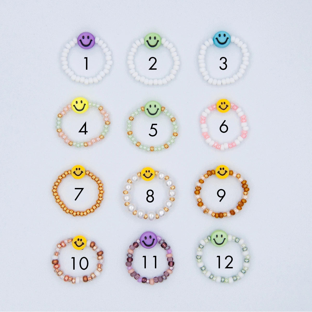 12 verschiedene Varianten von Smiley Fingerringen aus bunten Glasperlen mit je einer Smiley Perle auf hellgrauem Hintergrund
