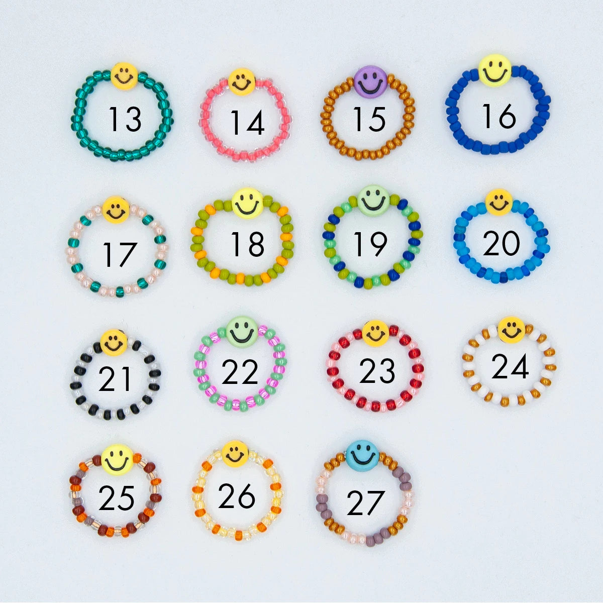 15 verschiedene Varianten von Smiley Fingerringen aus bunten Glasperlen mit je einer Smiley Perle auf hellgrauem Hintergrund