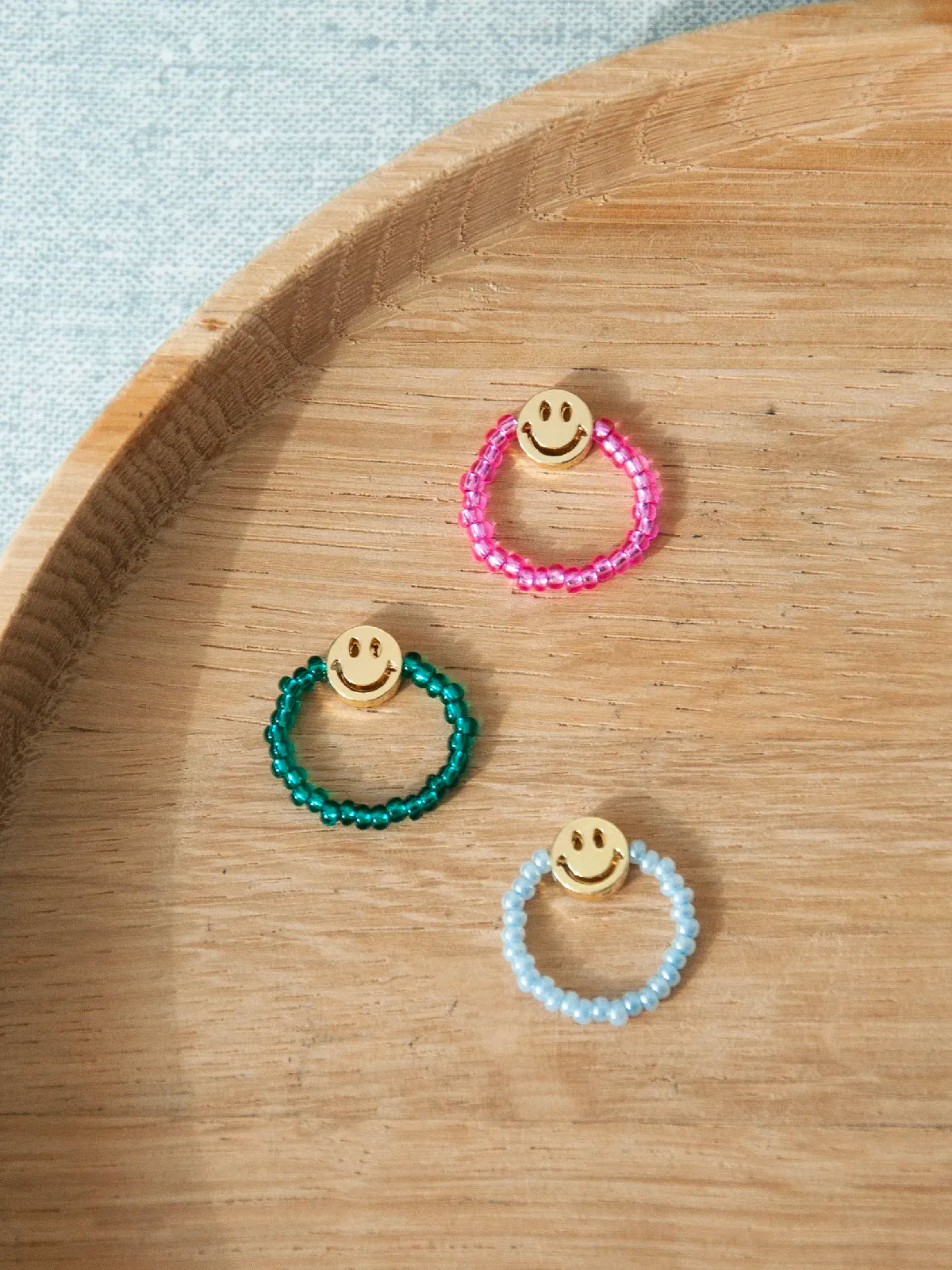 Drei bunte Fingerringe aus Glasperlen mit je einer goldigen Smielyperle auf holzigem Untergrund. Die Ringe sind pink, dunkelgrün und hellblau.