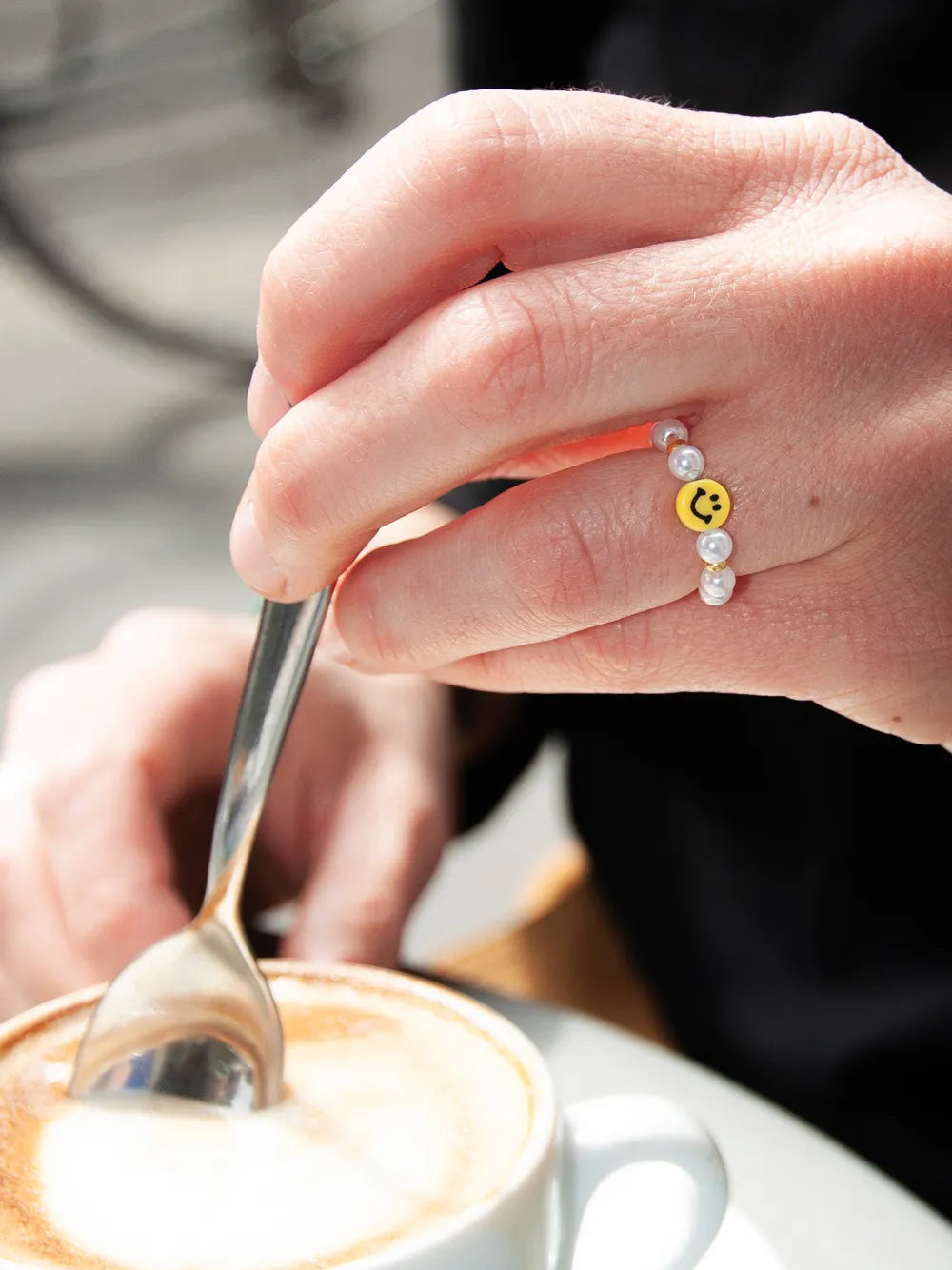 Eine Hand mit Smiley Fingerring hält Löffel und rührt in Cappucino