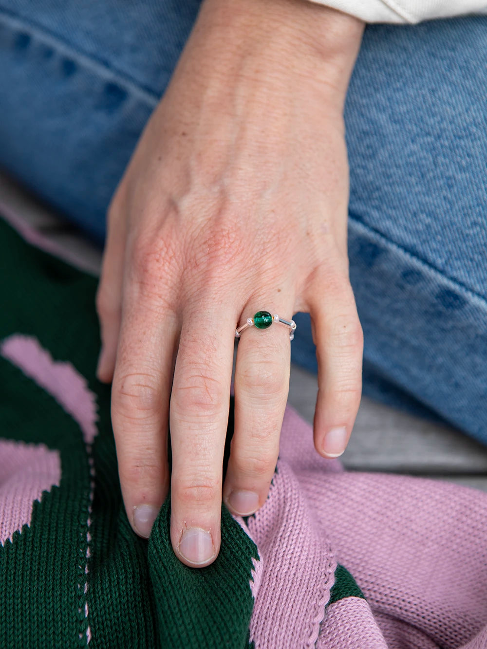 Detailansicht einer Hand welche einen Ring aus unterschiedlichen Glasperlen in grün-rosa trägt. Im Hintergrund sieht man einen Ausscnitt einer Jeans und eines grün-rosa farbenen Strickschals.