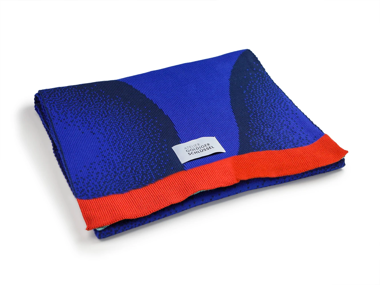 Gemusterter Jaquard Schal aus zwei unterschiedlichen Blautönen mit rot-orange farbenem Abschluss lieg gefaltet auf weissem Untergrund.