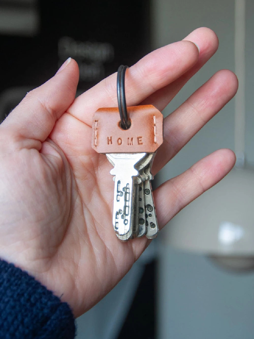 Hand hält Schlüsselbund, über vorderstem Schlüssel ein Schlüsselcover aus braunem Leder mit Prägung Home