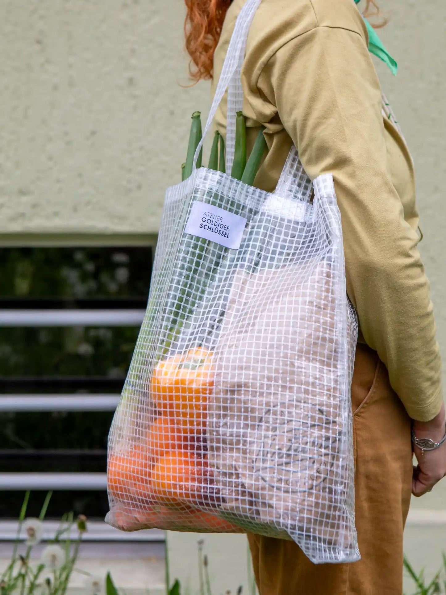 Eine Person trägt einen Shopper aus einem Kunststoffmaterial mit Gittermuster über der Schulter. Die Tasche ist gefüllt mit Peperonis, Orangen und Lauch. Im Hintergrund sieht man eine gelbgrüne Wand.