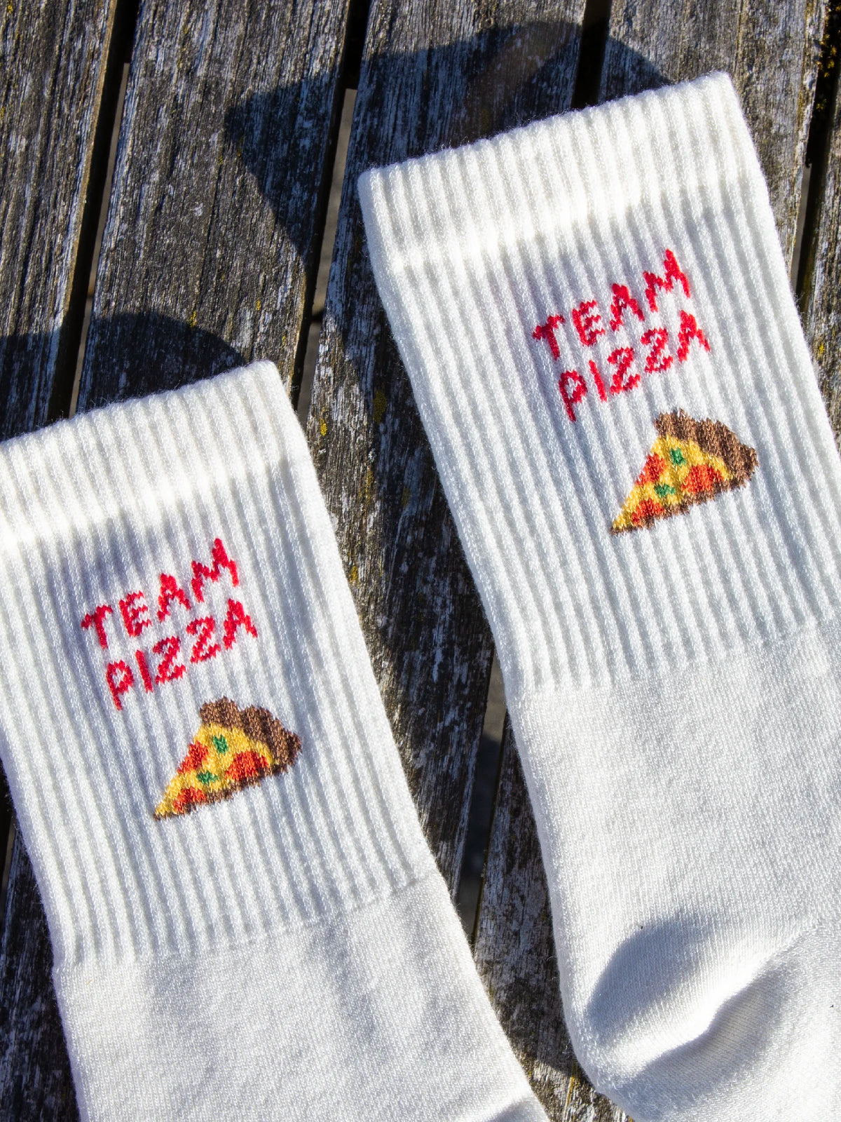 Detailansicht zweier ecru farbener Sportsocken welche im Bündchen den roten Schriftzug Team Pizza sowie ein Pizza Stück eingestrickt haben.