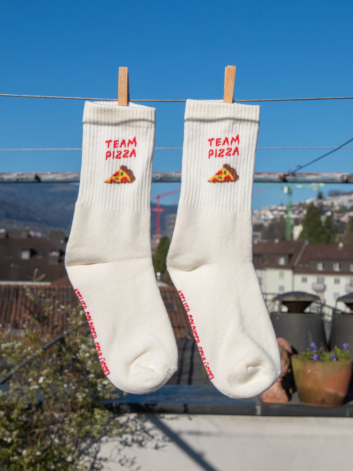 Zwei ecru farbenen Sportsocken hängen an einer Wäscheleine. Im Hintergrund sieht man unscharf Häuser, Hügel und einen blauen Himmel. Im Bündchen der beiden Socken ist der Schriftzug Team Pizza sowie ein Stück Pizza gestrickt.