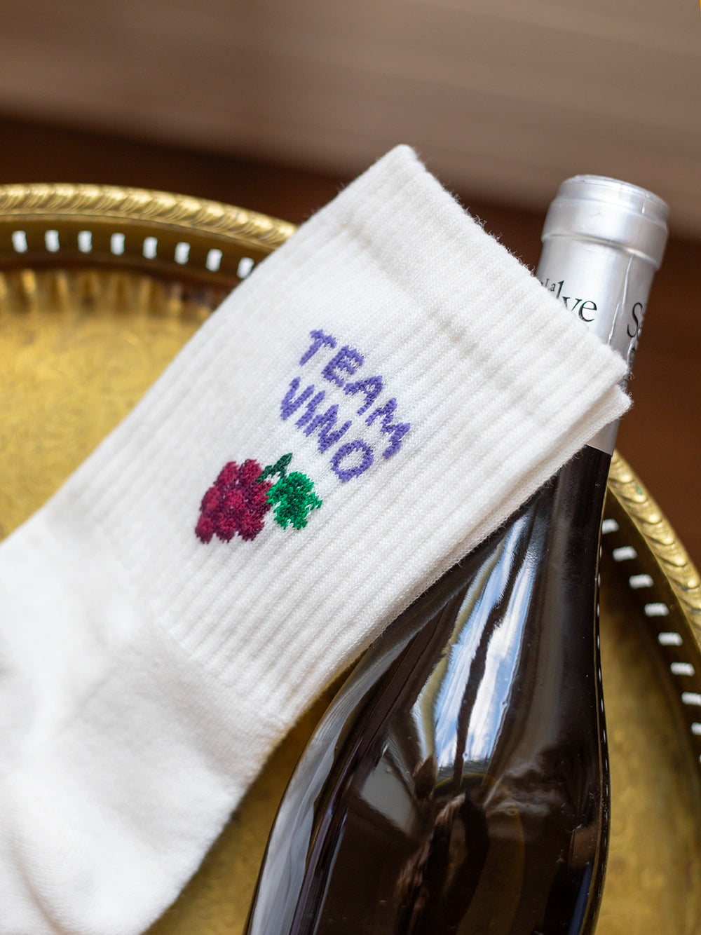 Anschnitt von weissen Sportsocken mit dem violetten Schriftzug «Team Vino» und darunter einer kleinen Abbildung einer Traube. Darunter liegt eine Flasche Wein auf eingem goldigen Tablett.
