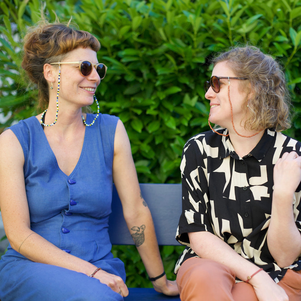 Miriam Schlüssel und Nina Goldiger sitzend auf Bank mit Sonnenbrillen und Brillenband und grün im Hintergrund.