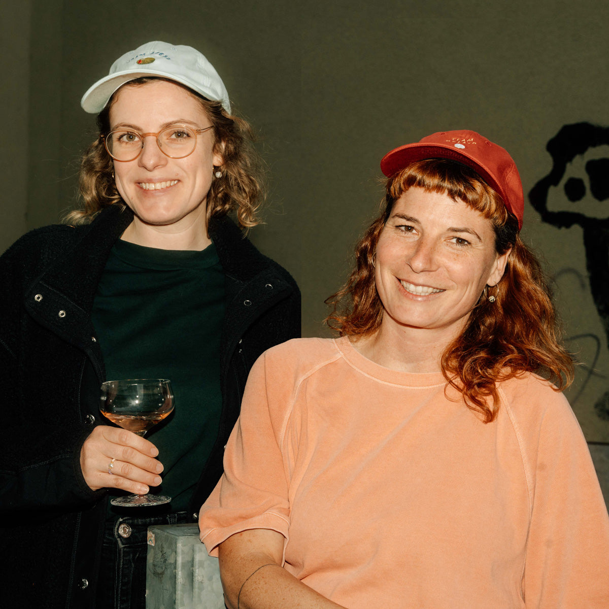 Nina Goldiger und Miriam Schlüssel mit bestickten Caps und Sektglas in der Hand