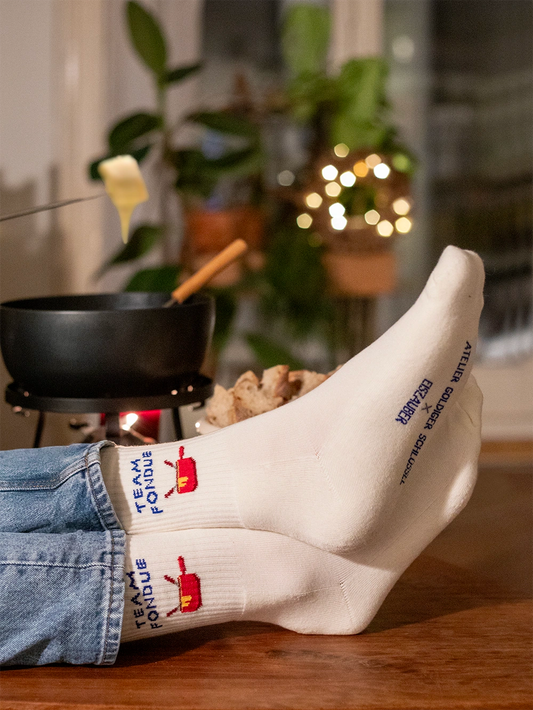 Detailausschnitt von Füssen die weisse Sportsocken mit dem Schriftzug Team Fondue tragen. Im Hintergrund unscharf ein Fondue Caquelon, eine Gabel mit Brot und geschmolzenem Käse daran und Lichter.