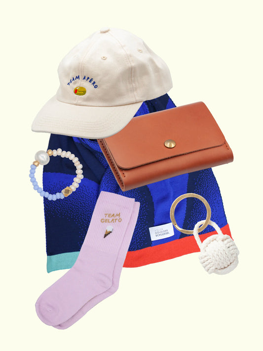 Collage von: weissem Team Apéro Cap, hellblau-beigem Glasperlenring mit Süsswasserperle, braunem Lederportemonnaie, hellrosa Team Gelato Socken, geknüpftem Schlüsselanhänger in weiss und blau gemustertem Schal