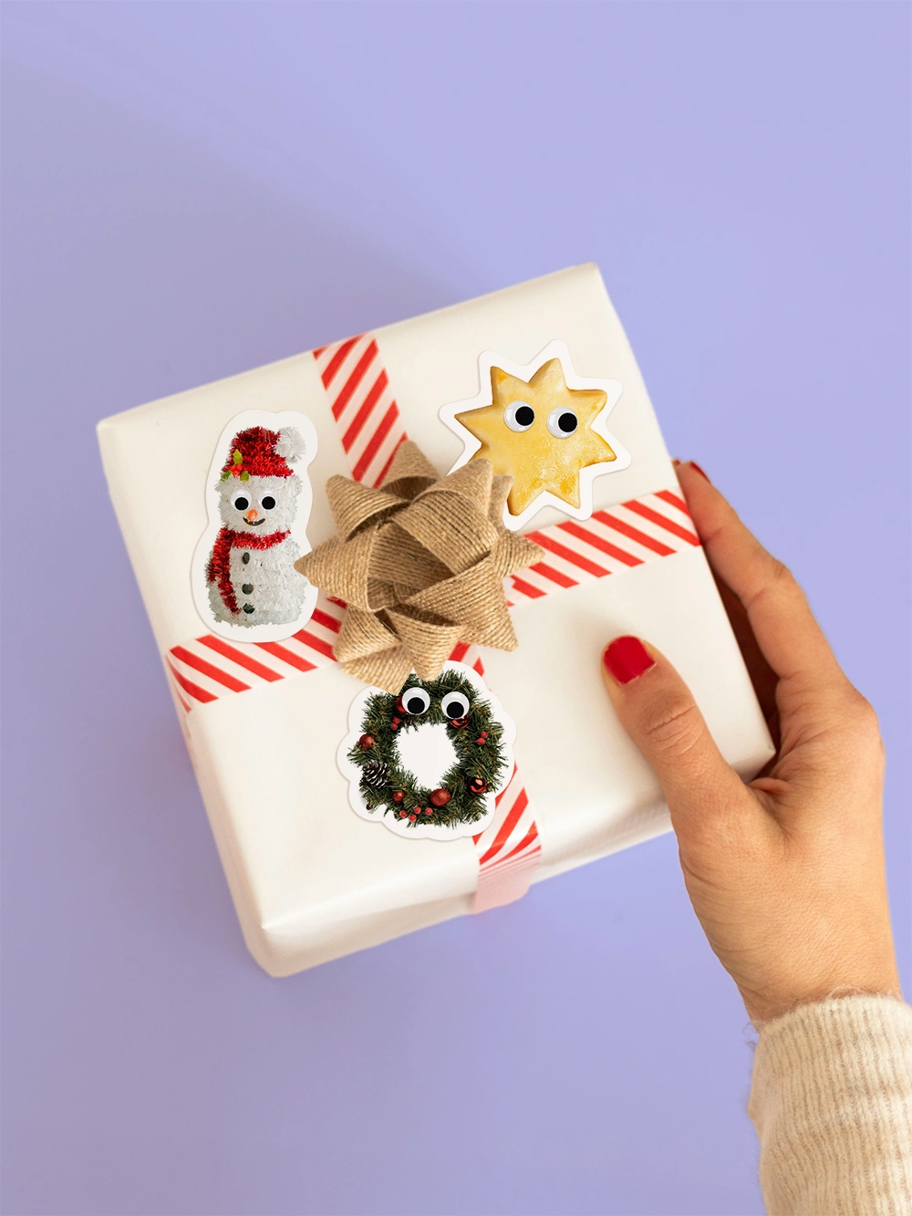 Aufsicht auf eine Hand die ein Geschenk mit gestreiftem Geschenkband hält. Das Geschenk ist dekoriert mit drei Stickern wie ein Mailänderli, ein Adventskranz und ein Schnee mann mit Wackleaugen.