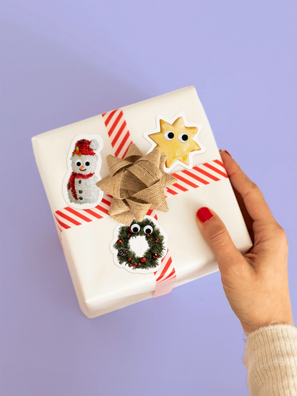 Aufsicht auf eine Hand die ein Geschenk mit gestreiftem Geschenkband hält. Das Geschenk ist dekoriert mit drei Stickern wie ein Mailänderli, ein Adventskranz und ein Schnee mann mit Wackleaugen.