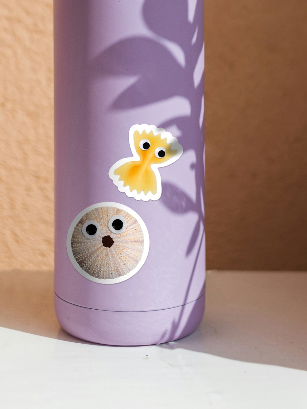 Ansicht einer lila Trinkflasche mit zwei Stickern in Form einer Farfalle Pasta und eines Seeigels mit Wackelaugen.
