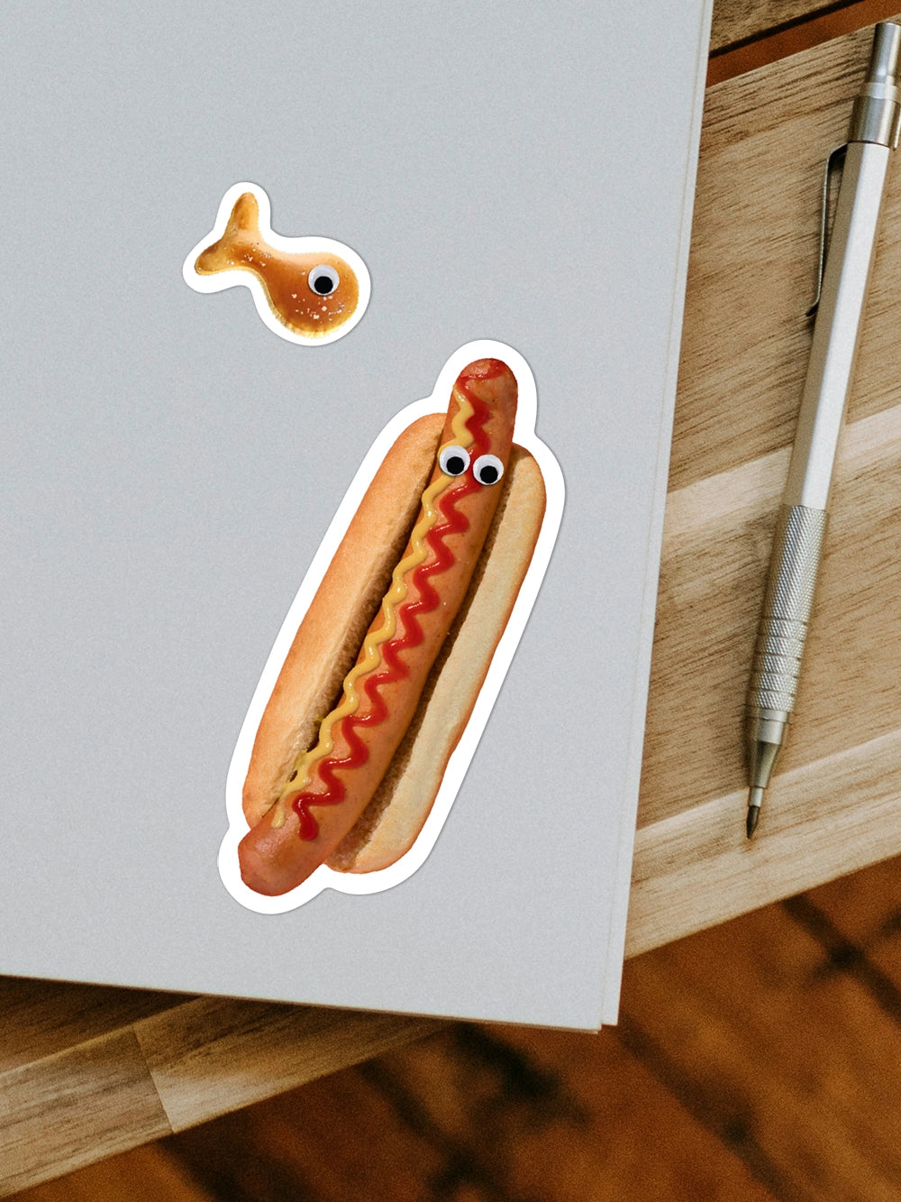 Aufsicht auf ein Notizheft mit zwei Stickern in Form eines Hotdogs mit Augen und eines Kambly Snackfischs. Nebenan liegt ein silberner Stift.