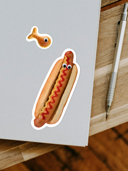 Aufsicht auf ein Notizheft mit zwei Stickern in Form eines Hotdogs mit Augen und eines Kambly Snackfischs. Nebenan liegt ein silberner Stift.